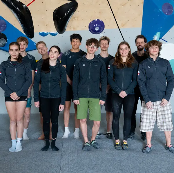 Een groep van tien jongvolwassenen staat binnen op een grijze mat; ze dragen allemaal dezelfde donkere jassen. Op de achtergrond is een klimwand te zien met diverse grepen en geometrische vormen. Een groep van tien jongvolwassenen staat binnen op een grijze mat; ze dragen allemaal dezelfde donkere jassen. Op de achtergrond is een klimwand te zien met diverse grepen en geometrische vormen.